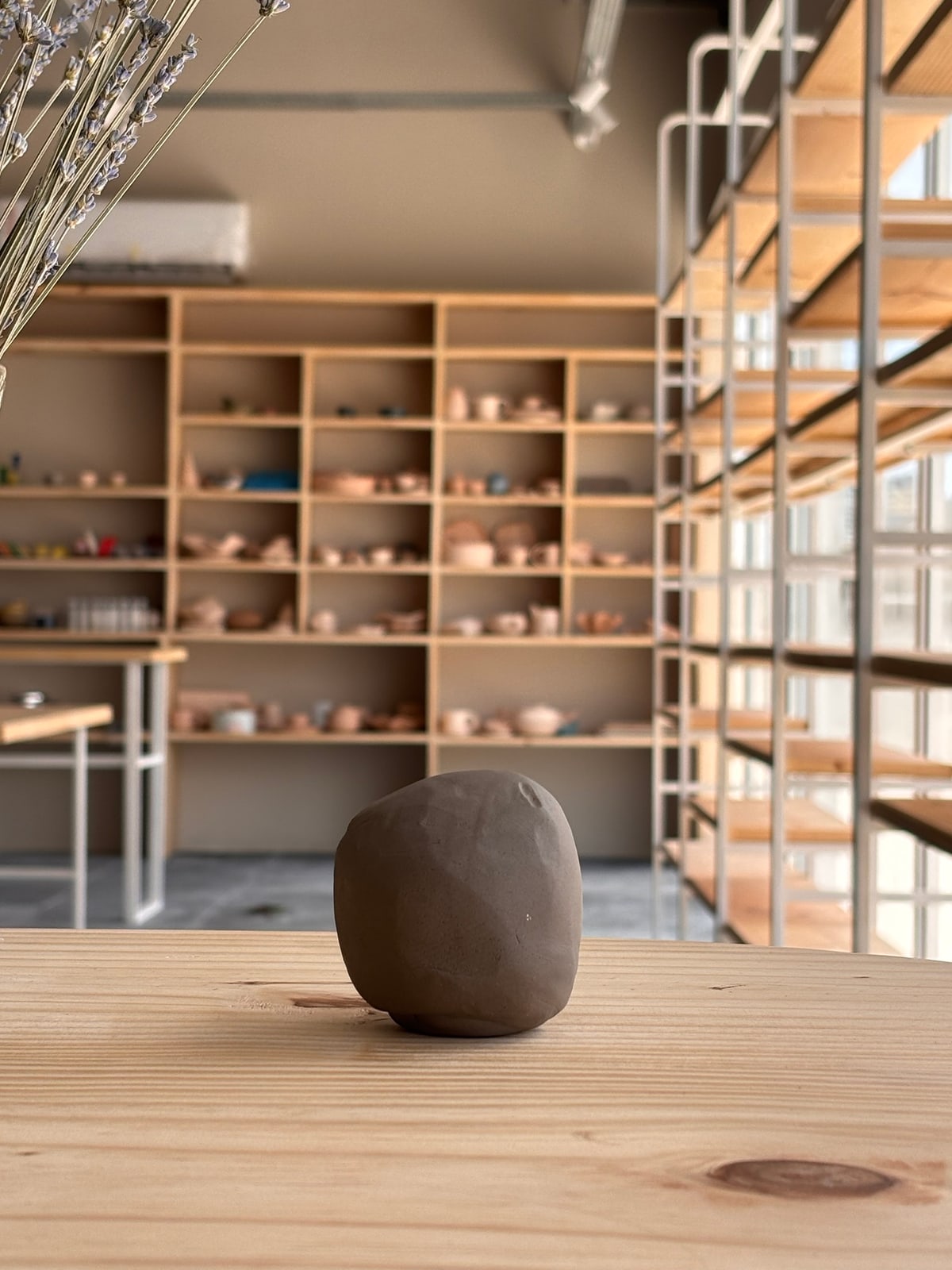Uma bola de argila crua sobre a mesa de madeira do ateliê, com as estantes de peças ao fundo